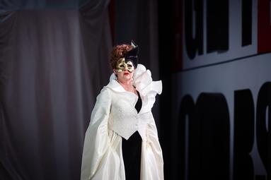 Vergrößerte Ansicht von Eine Person trägt ein auffälliges, weißes Kleid mit großen, voluminösen Ärmeln und einem strukturierten, asymmetrischen Oberteil. Das Kleid hat einen hohen Kragen und eine schwarze Kopfbedeckung mit einer geschwungenen Form. Der Hintergrund ist…