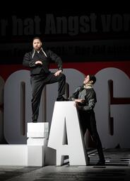 Vergrößerte Ansicht von Zwei Personen in schwarzer Kleidung, eine steht mit einem Bein auf einem weißen Podest, die andere lehnt an einem großen weißen Buchstaben A.