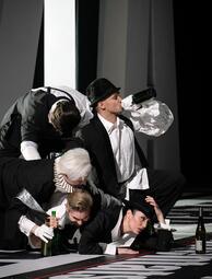 Vergrößerte Ansicht von Vier Personen in schwarz-weißer Kleidung liegen und knien auf einer schwarz-weißen Bühne, zwei halten grüne Flaschen, eine trinkt aus einer Flasche.