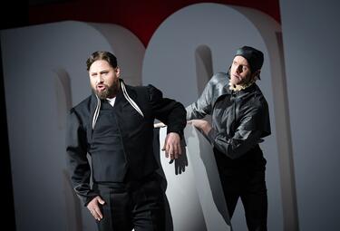 Vergrößerte Ansicht von Zwei Personen in schwarzer Kleidung lehnen an einem weißen Buchstaben vor einem dunklen Hintergrund.