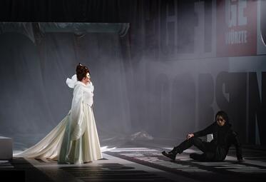 Vergrößerte Ansicht von Eine Frau in einem langen, weißen Kleid mit weitem Rock steht in einem dunklen Raum, ein Mann in schwarzer Kleidung sitzt auf dem Boden gegenüber.