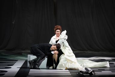 Vergrößerte Ansicht von Ein Mann in schwarzem Anzug lehnt mit geschlossenen Augen an den Knien einer sitzenden Frau in weißem, langem Kleid mit Rüschen auf der Schulter, vor schwarzem Hintergrund.