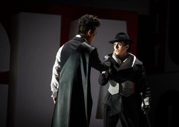 Vergrößerte Ansicht von Zwei Männer stehen sich gegenüber, einer mit einem langen schwarzen Umhang und weißem Hemd, der andere trägt einen schwarzen Hut, einen schwarz-weißen Anzug mit Weste und hält einen Gehstock. Die Szene wirkt theatralisch, mit dunklem Hintergrund und…