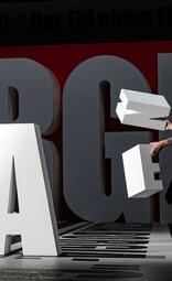 Vergrößerte Ansicht von Große, weiße Buchstaben stehen auf einem schwarz-weißen Boden mit einem rot-schwarzen Hintergrund. Eine Person hält zwei weiße Buchstaben, die sie gerade in die Luft hebt oder bewegt. Die Buchstaben sind massiv und dreidimensional, die Person ist…