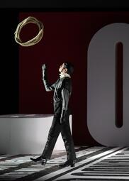 Vergrößerte Ansicht von Eine Person in schwarzer Kleidung, einschließlich Handschuhen und Schuhen, steht auf einem schwarz-weißen Boden mit Schrift. Die Person blickt nach oben und hebt eine Hand, während ein Seil in der Luft über ihr schwebt. Im Hintergrund ist ein großes…