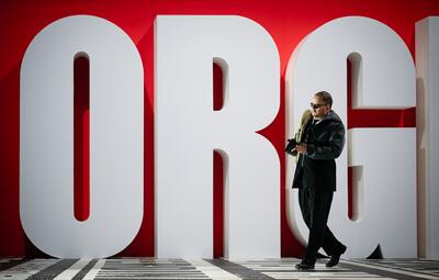 Vergrößerte Ansicht von Ein Mann in schwarzem Anzug und schwarzem Mantel steht vor einer großen roten Wand, auf der in weißen, überlebensgroßen Buchstaben das Wort &#039;ORGI&#039; zu lesen ist. Er hält seine Lederhandschuhe in den Händen und blickt nach unten. Der Boden ist schwarz…