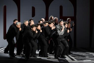 Vergrößerte Ansicht von Eine Gruppe von Personen steht eng beieinander auf einer Bühne mit dunklem Hintergrund und großen, teilweise sichtbaren Buchstaben in Weiß. Die Personen tragen überwiegend schwarze Kleidung, einer ein Oberteil in dunklen Grautönen. Sie sind in einer…