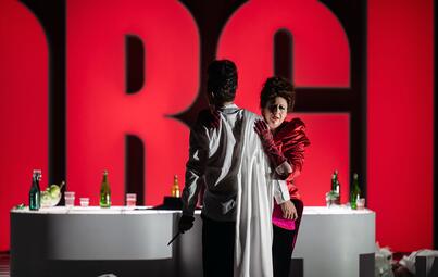 Vergrößerte Ansicht von Zwei Personen stehen vor einer großen, rot beleuchteten Wand mit den Buchstaben &quot;RG&quot;. Die Person links trägt ein weißes Hemd und hält in der rechten Hand einen Dolch. Die Person rechts trägt ein rotes Kleid. Im Hintergrund steht ein weißer Tisch mit…