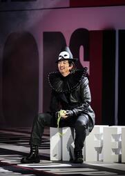 Vergrößerte Ansicht von Eine Person sitzt auf weißen, großen Buchstaben, die auf dem Boden angeordnet sind. Die Person trägt einen schwarzen Hut, eine schwarze Lederjacke und schwarze Hosen sowie schwarze Stiefel. Um den Hals trägt sie ein auffälliges, steifes, schwarzes…