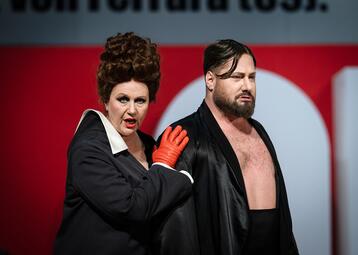 Vergrößerte Ansicht von Eine Person mit lockigem Haar trägt einen schwarzen Anzug mit weißem Hemd und orangefarbenen Handschuhen, die Hand auf der Schulter einer anderen Person ruhend. Die zweite Person trägt einen schwarzen, offenen Mantel, der die Brust freilegt. Im…