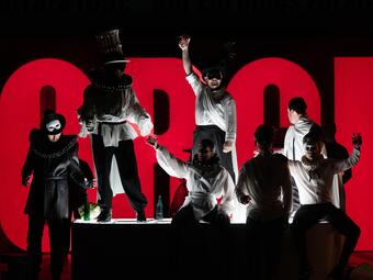 Vergrößerte Ansicht von Mehrere Personen stehen und sitzen vor einem großen, rot leuchtenden Schriftzug mit den Buchstaben &quot;ORGIA&quot;. Einige der Personen tragen weiße Hemden, eine Person trägt ein auffälliges Kostüm mit einem hohen Hut und einem großen, runden Kragen. Einige…