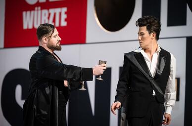 Vergrößerte Ansicht von Zwei Männer in schwarzer Kleidung stehen auf einer Bühne vor einem Hintergrund mit großen, teilweise sichtbaren Buchstaben in Schwarz und Rot. Der Mann links trägt einen langen schwarzen Mantel und hält einen silbernen Kelch mit ausgestrecktem Arm…
