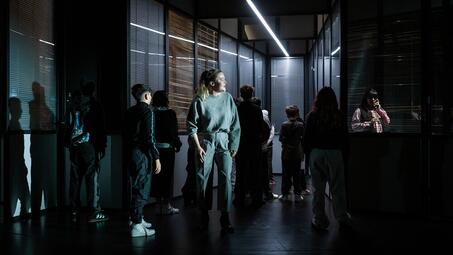 Vergrößerte Ansicht von Mehrere Personen stehen in einem dunklen Raum mit vertikalen Jalousien und einer langen Deckenleuchte, eine Person sitzt an einem Tisch mit Telefon.