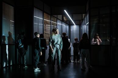 Vergrößerte Ansicht von In einem dunklen Raum stehen mehrere Personen, die meisten mit dem Rücken zur Kamera. Eine Person im Vordergrund trägt einen hellen Pullover und helle Hosen. Rechts im Bild sitzt eine Person an einem Tisch hinter einer Glaswand, die von einem…