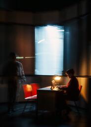 Vergrößerte Ansicht von In einem dunklen Raum sitzt eine Person an einem Schreibtisch, der von einer kleinen Lampe beleuchtet wird. Neben dem Schreibtisch stehen zwei rote Stühle. Im Hintergrund ist ein Fenster mit halb geschlossenen Jalousien, durch das helles Licht fällt…
