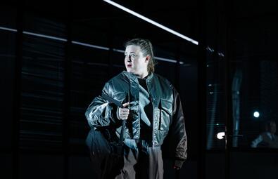 Vergrößerte Ansicht von Eine Person steht vor einem dunklen Hintergrund mit horizontalen Lichtstreifen. Sie trägt eine glänzende, schwarze Bomberjacke, darunter ein graues Oberteil, und hält eine schwarze Tasche über der Schulter. Die Person hat die Haare zu einem Zopf…