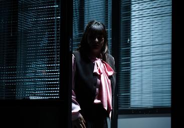 Vergrößerte Ansicht von Eine Person steht in einem dunklen Raum vor einem Fenster mit geschlossenen Jalousien. Die Person trägt eine schwarze Weste über einer hellrosa Bluse mit einer großen Schleife am Kragen. Die Arme hängen locker an den Seiten herunter. Das Licht fällt…