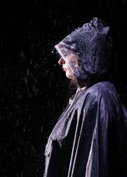 Vergrößerte Ansicht von Eine Person in einem dunklen, wasserabweisenden Regenmantel mit Kapuze steht im Regen. Der Mantel ist nass und glänzt durch die Regentropfen, die auf ihn fallen. Die Person ist seitlich zu sehen, der Kopf ist leicht nach oben geneigt.