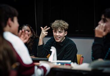 Vergrößerte Ansicht von In einem dunklen Raum sitzen mehrere Personen an einem Tisch. Eine Person mit kurzen, blonden Haaren trägt ein schwarzes Oberteil und hebt die rechte Hand mit zwei ausgestreckten Fingern. Auf dem Tisch stehen verschiedene Gegenstände, wie in einem…