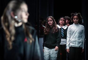 Vergrößerte Ansicht von Vier Jugendliche stehen in einer Reihe vor einem dunklen Hintergrund. Die vorderste Person trägt eine schwarze Kapuzenjacke und helle Hosen, die zweite Person ein weißes Hemd mit einem blauen und schwarzen gestreiften Krawattenmuster, die dritte…