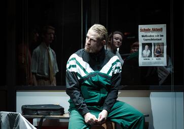 Vergrößerte Ansicht von Eine Person in einem grün-weißen Trainingsanzug sitzt auf einer Bank vor einer Glaswand. Hinter der Glaswand sind zwei weitere Personen zu sehen. An der Glaswand hängt ein Plakat mit der Überschrift 'Schule Aktuell' und dem Text 'Methoden wie im…