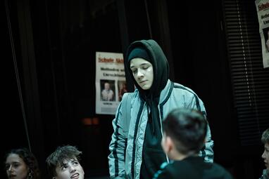 Vergrößerte Ansicht von Eine Person mit Kapuzenpullover und einer hellen, gesteppten Jacke steht in einem dunklen Raum. Im Hintergrund sind zwei Plakate an der Wand zu sehen, auf einem ist teilweise der Text 'Schule' und 'Methoden' lesbar. Vor der Person sind weitere…