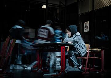 Vergrößerte Ansicht von Ein Schüler sitzt an einem Tisch in einem dunklen Raum, trägt eine graue Jacke mit Kapuze und schwarze Hosen mit weißen Streifen. Um ihn herum sind weitere Personen in Bewegung, deren Konturen verschwimmen, was auf Bewegung hindeutet. Der Tisch und…