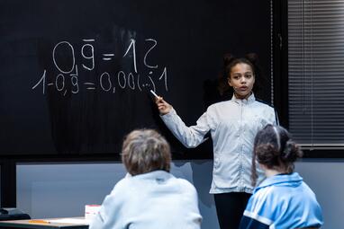 Vergrößerte Ansicht von Ein Kind steht vor einer Tafel und zeigt mit einem Zeigestock auf eine mathematische Gleichung, die mit Kreide geschrieben ist. Die Gleichung lautet: '0,9 = 1' und darunter '1 - 0,9 = 0,00... = 1'. Zwei Kinder sitzen mit dem Rücken zur Kamera und…