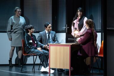 Vergrößerte Ansicht von In einem Raum mit geschlossenen Jalousien sitzen zwei Personen an einem Tisch, eine dritte Person steht dahinter. Die sitzenden Personen tragen formelle Kleidung, darunter ein Anzug und ein dunkelblauer Pullover. Am Tisch sitzt eine Frau mit langem…