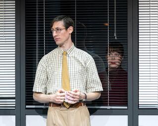 Vergrößerte Ansicht von Ein Mann steht vor einem Fenster mit weißen Jalousien. Er trägt ein kurzärmeliges, kariertes Hemd in Weiß und Beige, eine gelbe Krawatte sowie eine beige Hose mit braunem Gürtel. Er hält eine Tasse mit beiden Händen vor sich. Im Fenster sieht man…