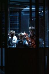 Vergrößerte Ansicht von Drei Personen stehen hinter einer Glaswand mit vertikalen und horizontalen Lamellen, die Lichtstreifen auf ihre Kleidung und Hände werfen. Zwei der Personen halten ihre Hände an die Scheibe, während die dritte Person seitlich steht. Die Szene ist…