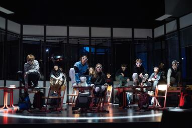 Vergrößerte Ansicht von In einem dunklen Raum mit großen Fenstern sitzen mehrere Kinder auf und an Schulstühlen und -tischen, die in einer Reihe angeordnet sind. Die Kinder tragen unterschiedliche Kleidung, darunter Pullover und Jacken. Einige Kinder sitzen auf den Tischen…