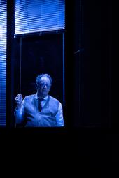 Vergrößerte Ansicht von Ein Mann steht hinter einem Fenster mit heruntergelassener Jalousie, die er mit der rechten Hand hält. Er trägt ein helles Hemd, eine dunkle Krawatte und eine helle Strickweste. Der Hintergrund ist dunkel, und das Bild ist in einem kühlen Blauton…