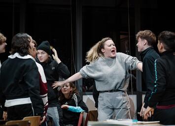 Vergrößerte Ansicht von Eine Gruppe von Jugendlichen steht in einem Raum mit großen Fenstern im Hintergrund. Eine Person in der Mitte trägt einen hellgrauen Pullover und hellgraue Hosen, die Arme sind seitlich ausgestreckt. Die anderen Jugendlichen tragen überwiegend…