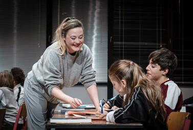 Vergrößerte Ansicht von Eine Frau in einem grauen Pullover und hellgrauer Hose beugt sich über einen Tisch, auf dem mehrere Bücher und Schreibmaterialien liegen. Vor ihr sitzen zwei Kinder, die an ihren Schreibtischen schreiben. Im Hintergrund sind weitere Kinder an…