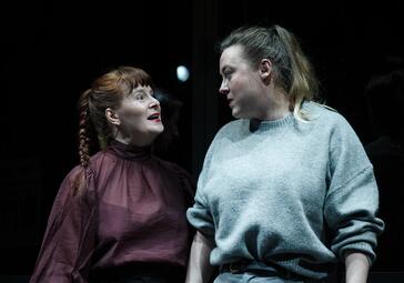 Vergrößerte Ansicht von Zwei Frauen stehen nebeneinander vor einem dunklen Hintergrund. Die Frau links trägt eine hochgeschlossene, langärmelige Bluse in dunklem Rot mit einem leichten Glanz und hat ihr rotbraunes Haar zu einem Zopf geflochten. Die Frau rechts trägt einen…