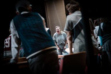 Vergrößerte Ansicht von In einem dunklen Raum steht eine Person in grauem Pullover vor einem Fenster mit geschlossenen Jalousien. Im Vordergrund sind mehrere Personen zu sehen, die sich zu der stehenden Person hinwenden. Die Personen im Vordergrund tragen unterschiedliche…