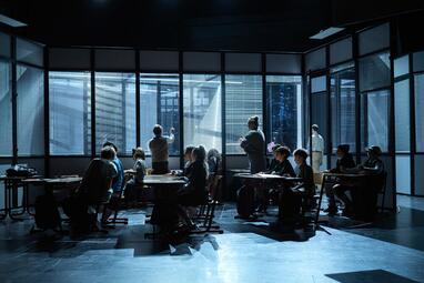 Vergrößerte Ansicht von In einem dunklen Raum sitzen mehrere Personen an Tischen, die in zwei Reihen angeordnet sind. Die Personen sind von der Seite oder von hinten zu sehen und wirken in eine Richtung, die von links nach rechts verläuft. Im Hintergrund sind große Fenster…