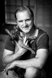 Vergrößerte Ansicht von ein schwarz-weiß Foto von Thommy Mardo. Er sitzt in der Hocke, lächelt in die Kamera und hat einen kleinen Hund auf dem Arm.