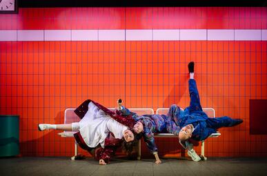 Vergrößerte Ansicht von Zwei Schauspieler liegen auf einer weißen Bank vor einer orangefarbenen Kachelwand, einer trägt ein buntes Hemd und blaue Hose, der andere ein weißes Kleid und roten Mantel.