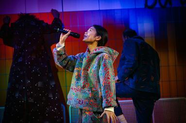 Vergrößerte Ansicht von Schauspielerin im bunten, gemusterten Kostüm hält ein Mikrofon und steht auf der Bühne.