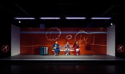 Vergrößerte Ansicht von Drei Schauspieler sitzen auf einer Bühne mit rotem Hintergrund, auf dem weiße und orange Linien eine Landschaft mit Wolken und einem Baum zeichnen.