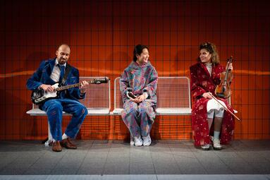 Vergrößerte Ansicht von Drei Personen sitzen auf einer weißen Bank vor orangefarbenen Kacheln, links ein Mann in blauem Anzug mit E-Gitarre, in der Mitte eine Person in buntem Pullover, rechts eine Frau in rotem Mantel mit Geige.