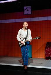 Vergrößerte Ansicht von Person mit weißem Hemd und blauer Hose mit buntem Muster spielt E-Gitarre vor orange-weiß gekachelter Wand mit Uhr im Theaterstück Mutter Dili am Jungen Nationaltheater Mannheim.