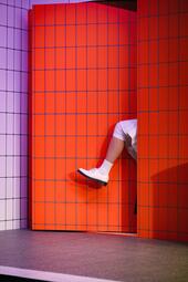 Vergrößerte Ansicht von Ein Bein mit weißem Schuh und weißer Socke ragt aus einer orange gefliesten Tür, die in eine weiß und orange geflieste Wand eingebettet ist.