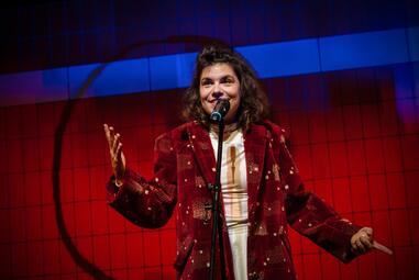 Vergrößerte Ansicht von Schauspieler mit langem Haar trägt roten Samtblazer mit goldfarbenen Mustern und steht vor rotem Hintergrund mit schwarzem Gittermuster, spricht in Mikrofon.