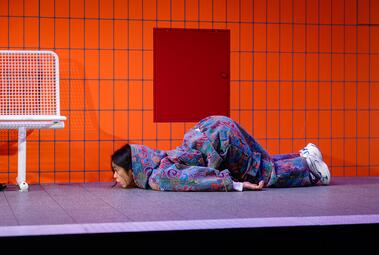 Vergrößerte Ansicht von Person liegt bäuchlings auf dem Boden vor einer orange gekachelten Wand mit rotem Schaltkasten, trägt bunte, gemusterte Kleidung und weiße Sportschuhe.