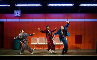 Vergrößerte Ansicht von Drei Personen in bunter Kleidung tanzen auf einer Bahnsteigbühne mit rotem Kachelhintergrund und weißer Sitzbank, darüber eine Uhr.