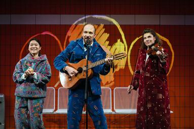 Vergrößerte Ansicht von Drei Personen auf der Bühne, eine spielt Gitarre, eine Geige, alle tragen bunte, gemusterte Kostüme vor rotem Hintergrund mit stilisiertem Gesicht.