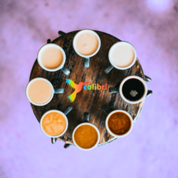 Vergrößerte Ansicht von Ein Blick von oben auf einen runden Tisch. Darauf stehen Tassen im Kreis, gefüllt mit unterschiedlich hellem und dunklem Kaffee. In der Mitte ein bunter Kolibri-Vogel neben dem Wort colibri.